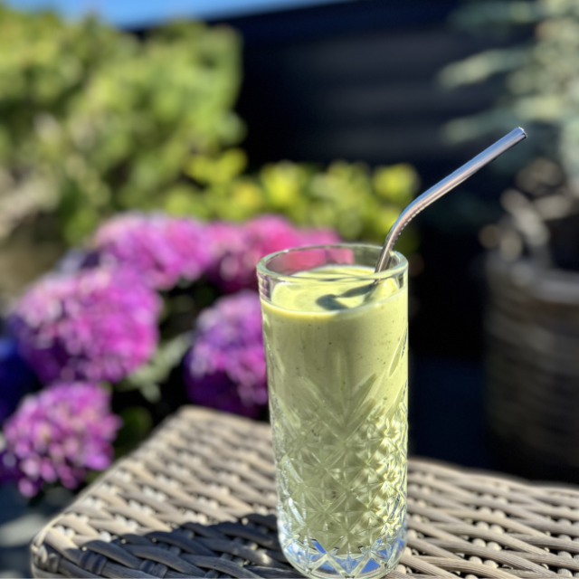Green avocadosmoothie in a glass with a straw on a wicker table outdoors, surrounded by flowers.
