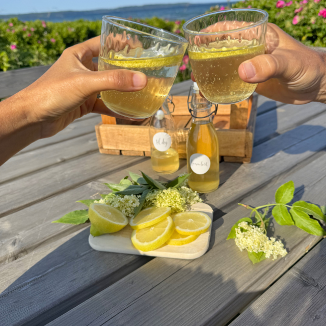Two people clinking glasses of lemonade on a wooden table with lemons and flowers.